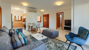 A modern living room with a grey sofa, a blue armchair, and a coffee table. There is a dining area with a table and chairs in the background. - 2 Bedroom Apartment - CB02 (kouklia)