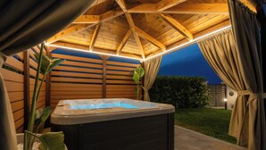 Ein Whirlpool in einer Außenanlage mit Holzbalken darüber und Blick auf ein Haus im Hintergrund.
