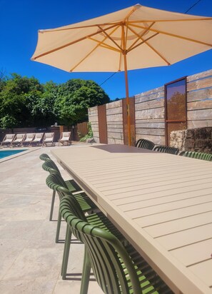 A poolside area with a long table and chairs, a large umbrella, and a wooden fence.