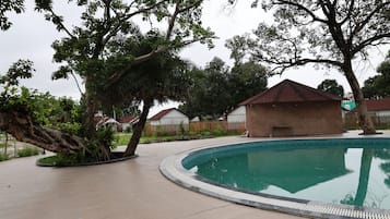 A swimming pool with a curved edge, surrounded by a paved walkway. There are trees and a building with a red roof in the background.