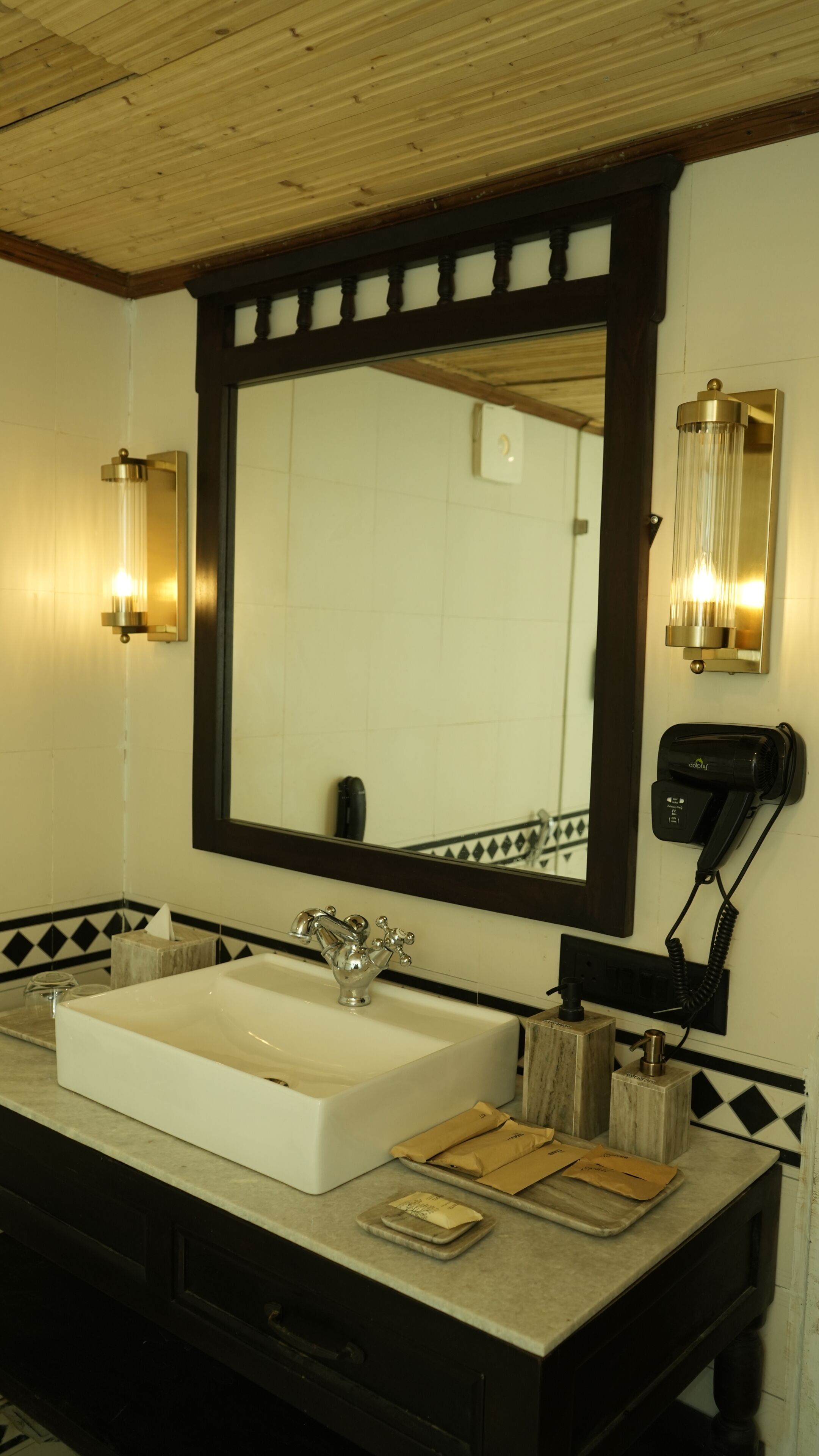 A bathroom with a large mirror, a white sink, two wall-mounted lights, a towel dispenser, and a soap dispenser.