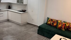 A modern kitchen with white cabinets and a marble countertop, a green sofa with colorful cushions, and a patterned rug.
