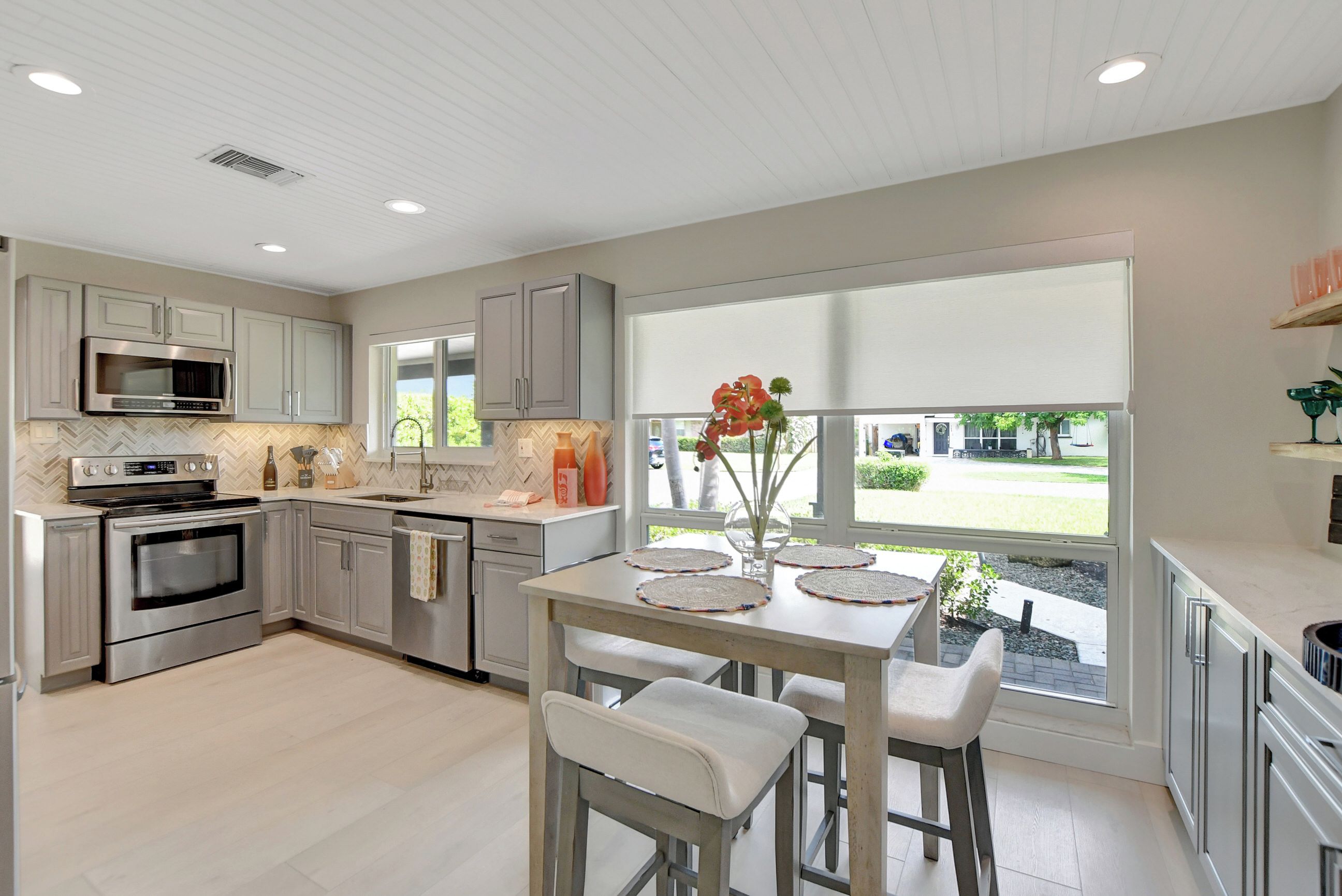A modern kitchen with stainless steel appliances, a dining area with a table and bar stools, and a large window offering a view of the outdoors.