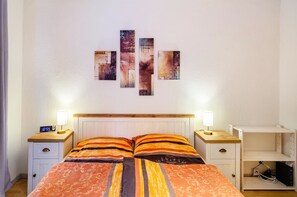 Un dormitorio con una cama, dos mesitas de noche, una mesita auxiliar, un estante en la pared y una obra de arte colgada.