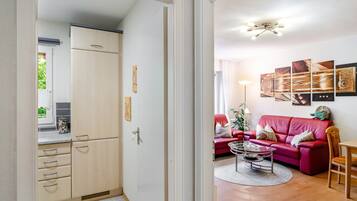 Ein Zimmer mit einer roten Couch, einem Couchtisch und einem Esstisch. Es gibt eine Küchenzeile mit Schränken und einem Kühlschrank.
