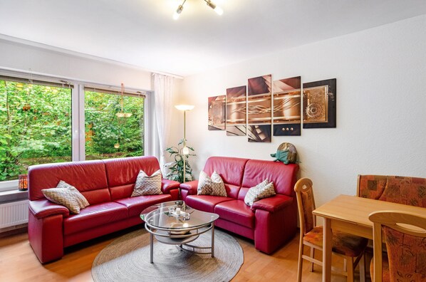 Una sala de estar con un sofá rojo, una mesa de centro de vidrio y una mesa de comedor con sillas. Hay obras de arte enmarcadas en la pared y un ventanal con vistas a zonas verdes.