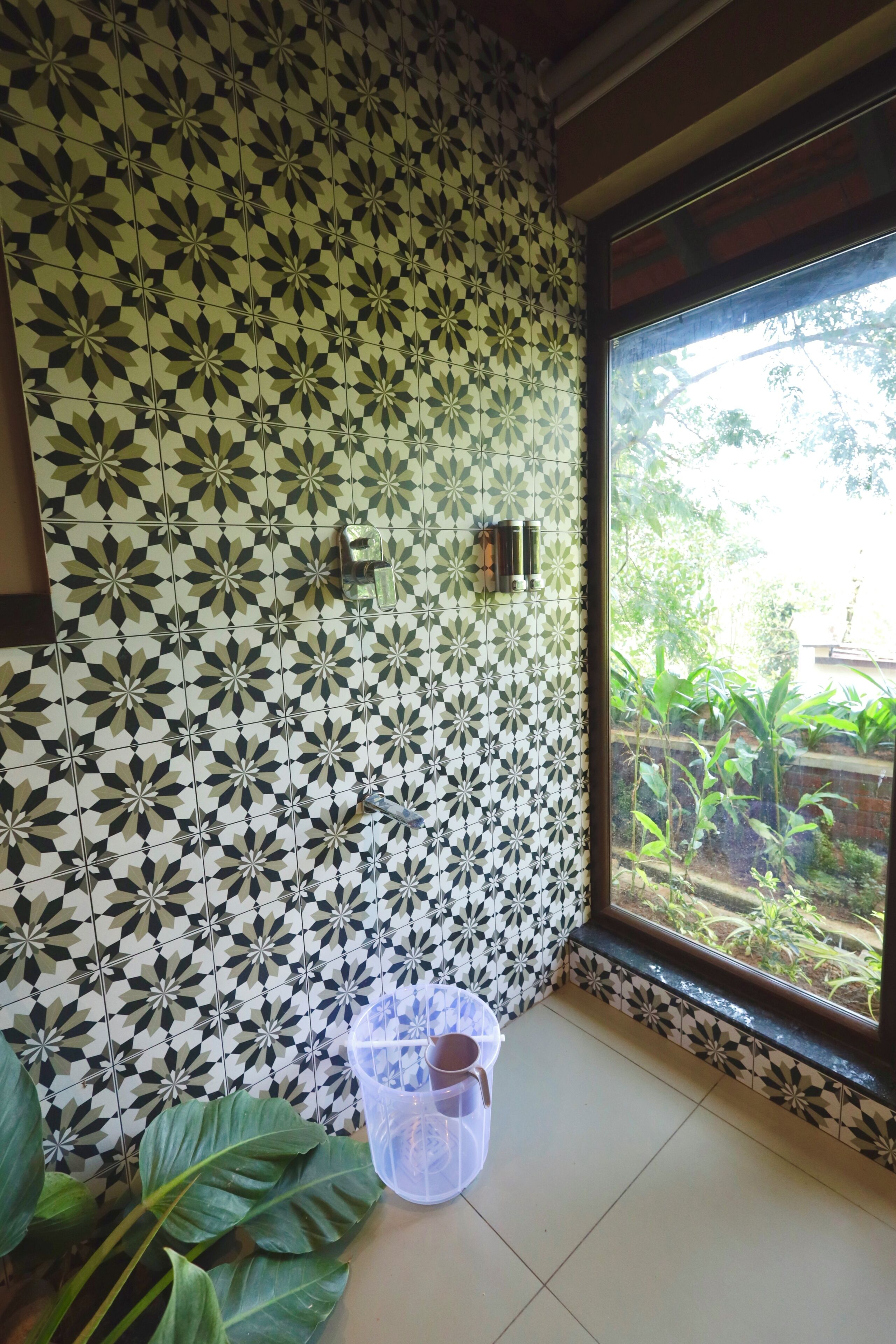 A bathroom with patterned tiles, a window with a view of greenery, and a plastic container on the floor.
