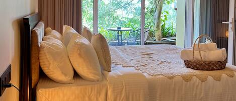A bedroom with a large window, a bed with white linens, a bedside lamp, and a wicker basket with towels.
