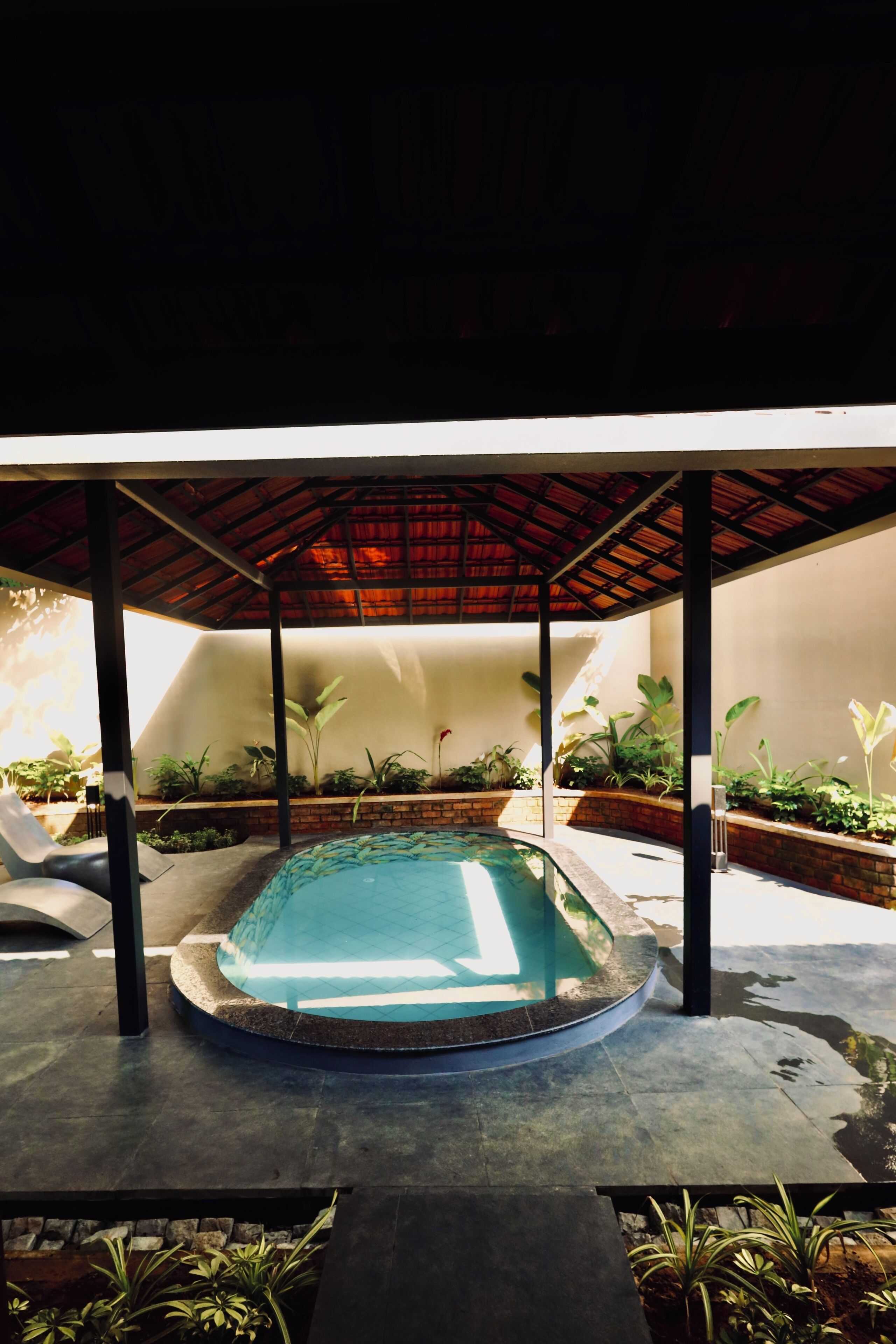 A covered swimming pool with a curved edge, surrounded by potted plants and a brick wall.