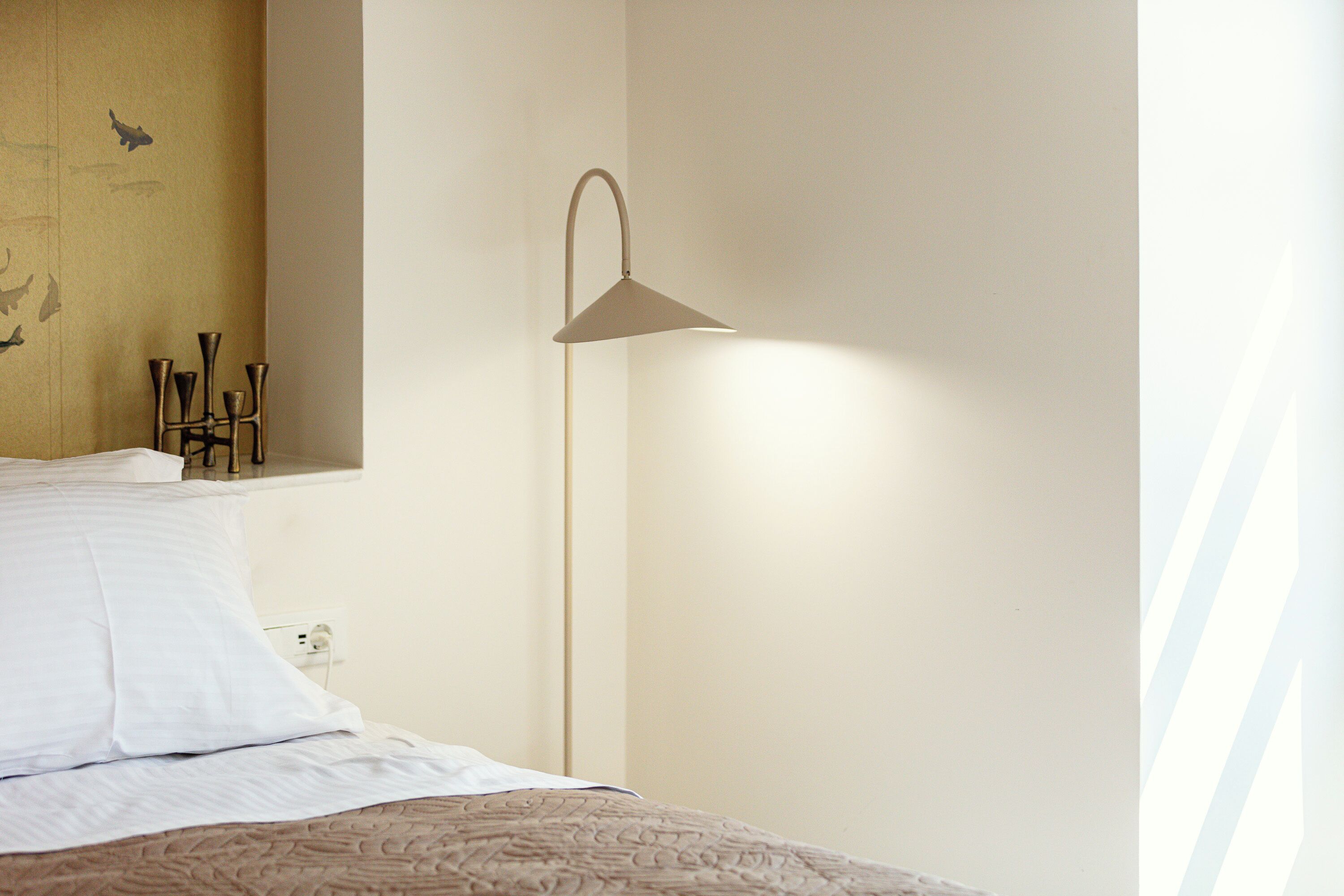 Una cama bien hecha con sábanas blancas, una mesita de noche con lámpara y una pared con diseño sencillo.