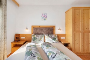 A bedroom with a wooden headboard, two bedside tables, a bed with leaf-patterned bedding, a wooden wardrobe, and a wall-mounted light fixture.