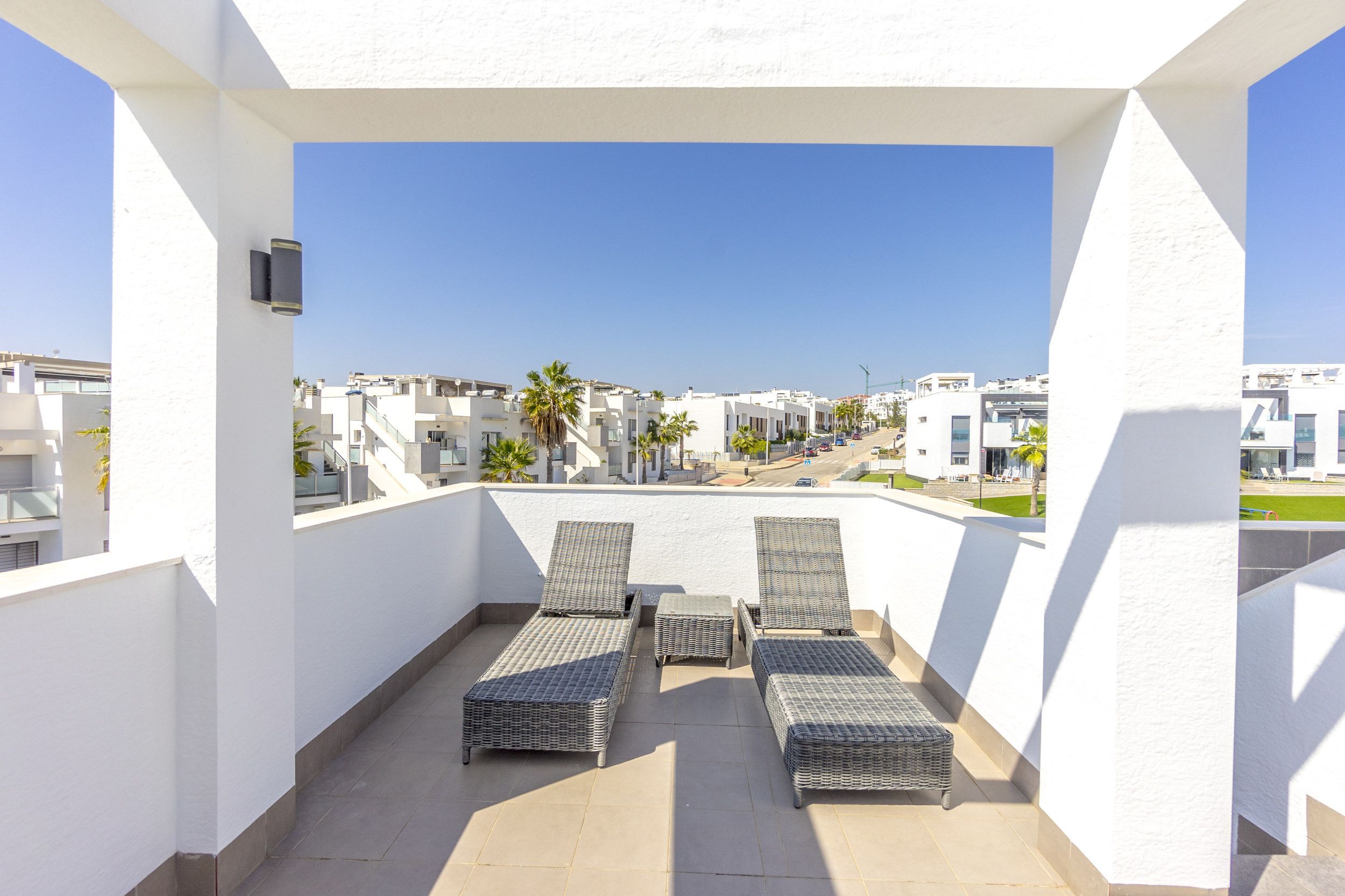 Un balcon agrémenté de quatre fauteuils de jardin en osier et d’une petite table, offrant une vue sur un quartier résidentiel avec des bâtiments blancs et des palmiers.