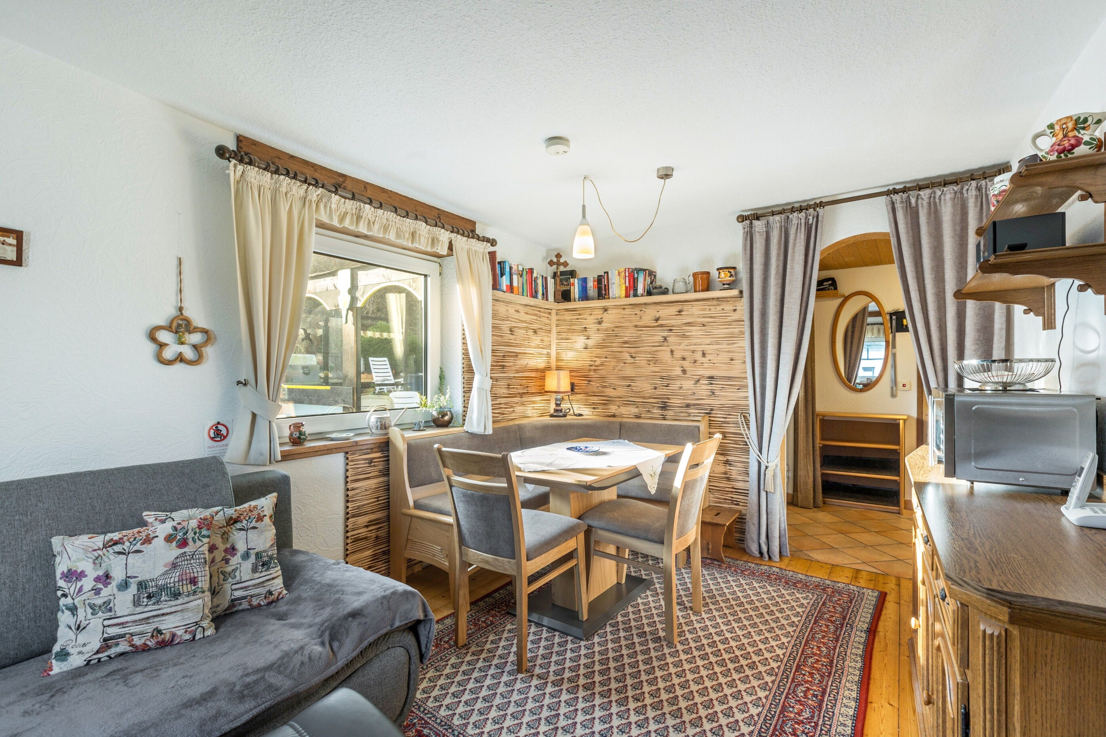 Ein gemütliches Zimmer mit einer Couch, einem Esstisch mit Stühlen, einem Bücherregal, einer kleinen Küchenzeile und einem Fenster mit Vorhängen.