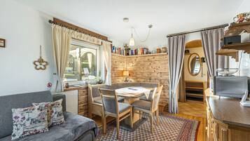 Ein gemütliches Zimmer mit einer Couch, einem Esstisch mit Stühlen, einem Bücherregal, einer kleinen Küchenzeile und einem Fenster mit Vorhängen.