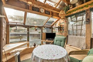 Een gezellige kamer met houten inrichting, een ronde tafel met een tafelkleed met bloemenprint, een groene fauteuil en een televisie.