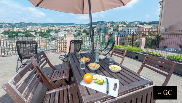 A terrace with a table set for dining, a large umbrella, and a city view in the background. - Elegant Apartment with a Breathtaking View over the Renaissance Genoa. (Genova)