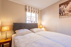 A double bed with a grey upholstered headboard, two bedside lamps, and a window with striped valance. - Käpt'n 25 - Käpt'n 25 Westerland (Sylt)
