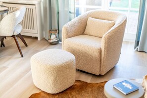 A cozy living room with a plush armchair and ottoman, a small table with a book, and a view of the outdoors through large windows. - Käpt'n 25 - Käpt'n 25 Westerland (Sylt)