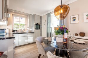 A modern kitchen with stainless steel appliances, a dining area with a round glass table, and a large window providing natural light. - Käpt'n 25 - Käpt'n 25 Westerland (Sylt)