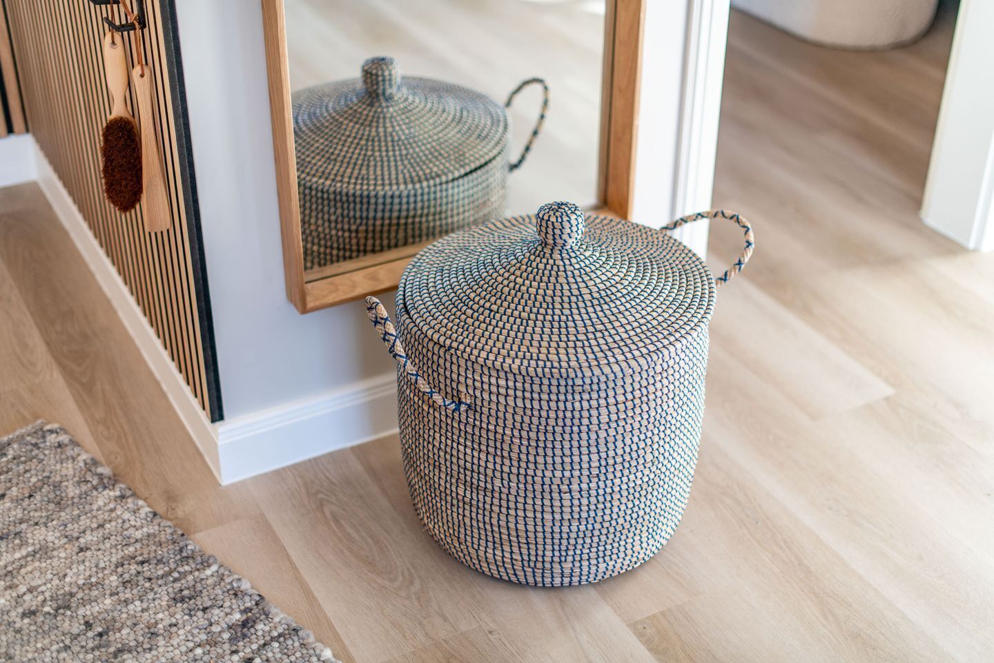 Un pouf rotondo intrecciato con maniglie, posizionato su un pavimento di legno vicino a uno specchio che riflette una cucina.