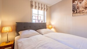 A double bed with a grey upholstered headboard, two bedside lamps, and a window with striped valance. - Käpt'n 25 - Käpt'n 25 Westerland (Sylt)