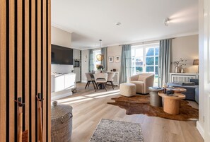 A modern living room with a sofa, ottoman, and coffee table, a dining area with chairs and a table, and a kitchen with a TV and appliances. - Käpt'n 25 - Käpt'n 25 Westerland (Sylt)