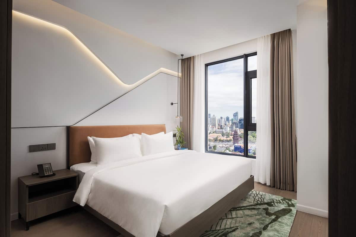 Quarto de hotel moderno com uma cama grande, mesa de cabeceira, luminária e vista para o horizonte urbano através de uma ampla janela.