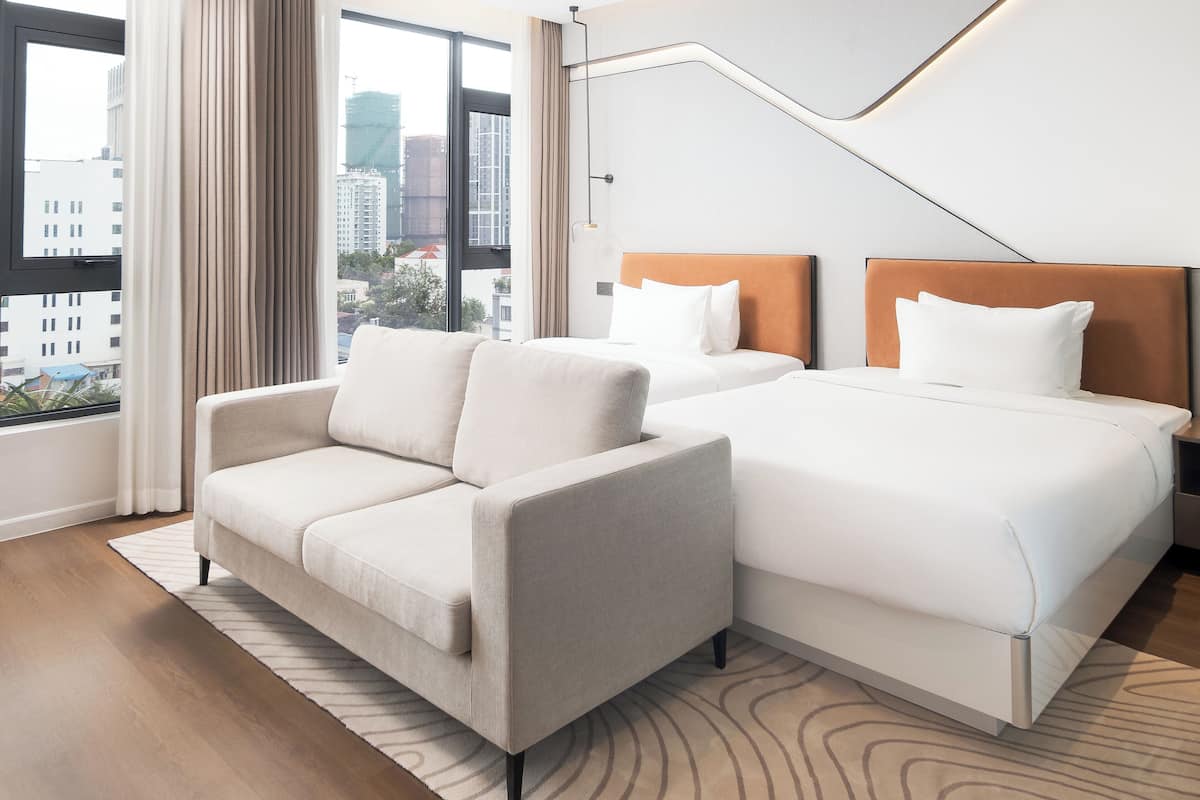 Quarto de hotel com duas camas, um sofá e uma janela ampla com vista para a cidade.