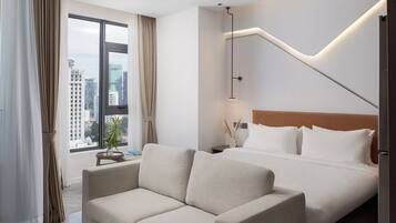 Habitación de hotel con cama, sofá, mesita de noche, lámpara y vista a la ciudad a través de un ventanal grande.