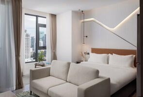 Habitación de hotel con cama, sofá, mesita de noche, lámpara y vista a la ciudad a través de un ventanal grande.