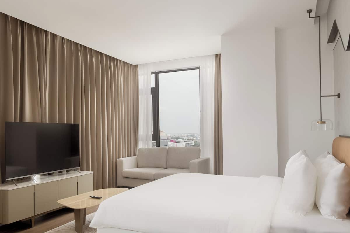 Quarto de hotel moderno com uma cama grande, uma televisão em um suporte, uma cadeira e vista para a cidade pela janela.