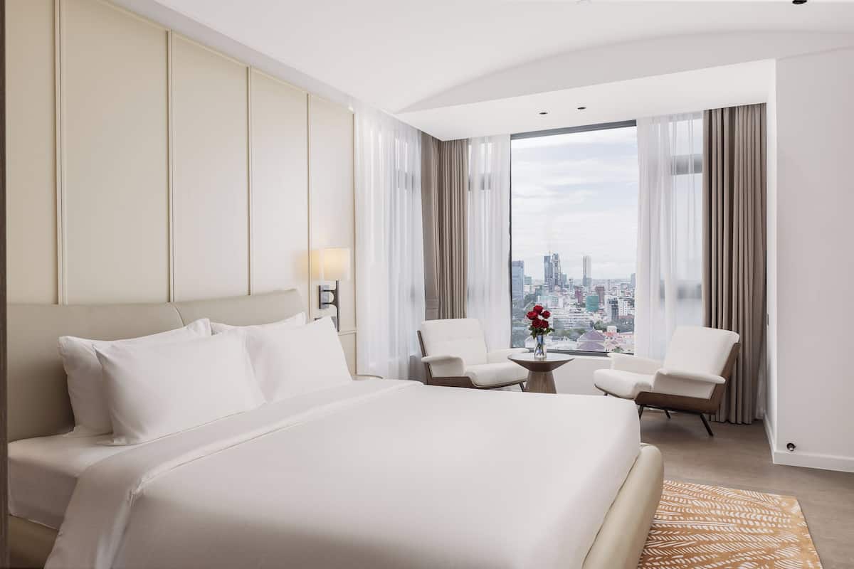 Quarto de hotel moderno com uma cama grande, duas poltronas, uma mesinha de centro com um vaso de flores e vista para a cidade através de amplas janelas.
