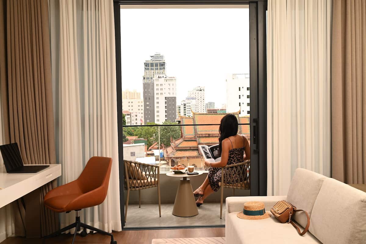 Quarto moderno com varanda, um laptop numa mesa, uma mesa de jantar com uma cadeira e um sofá com um chapéu e uma bolsa.