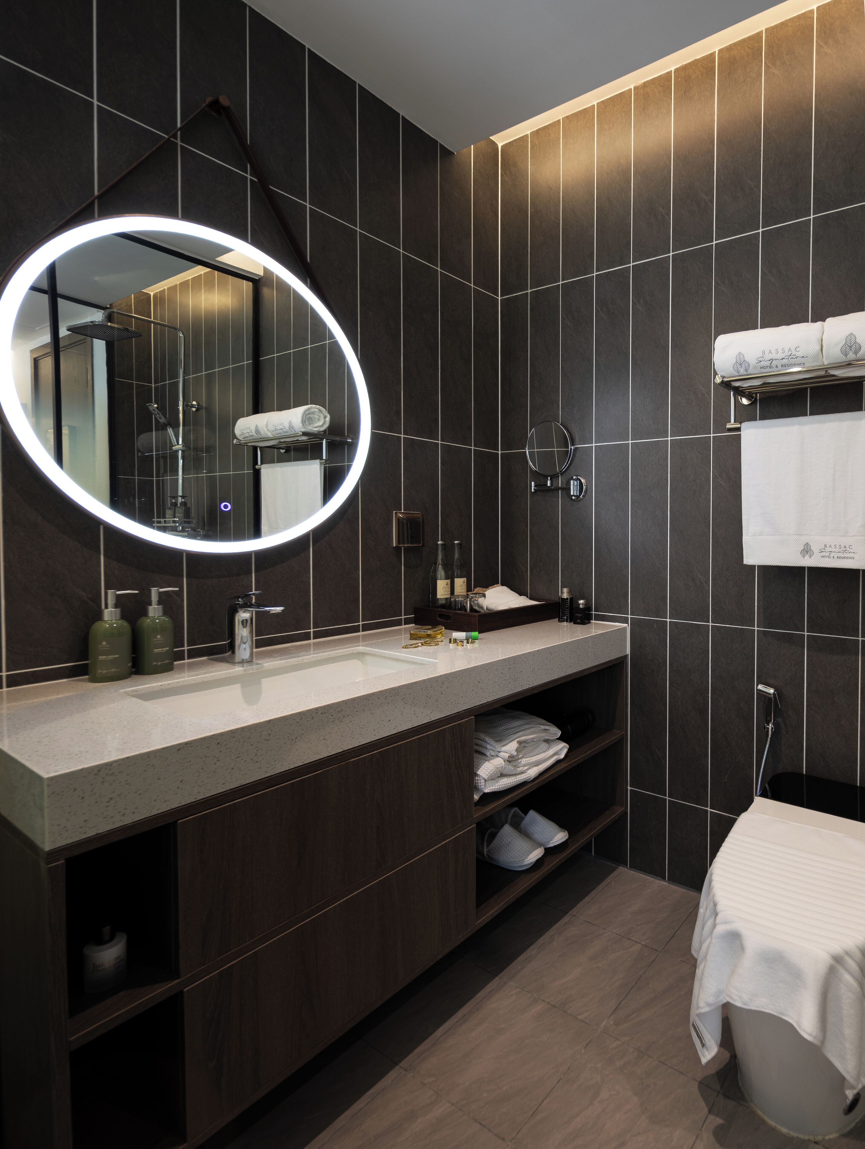 A modern bathroom with a large mirror, dark tiles, a sink with toiletries, and a towel rack.