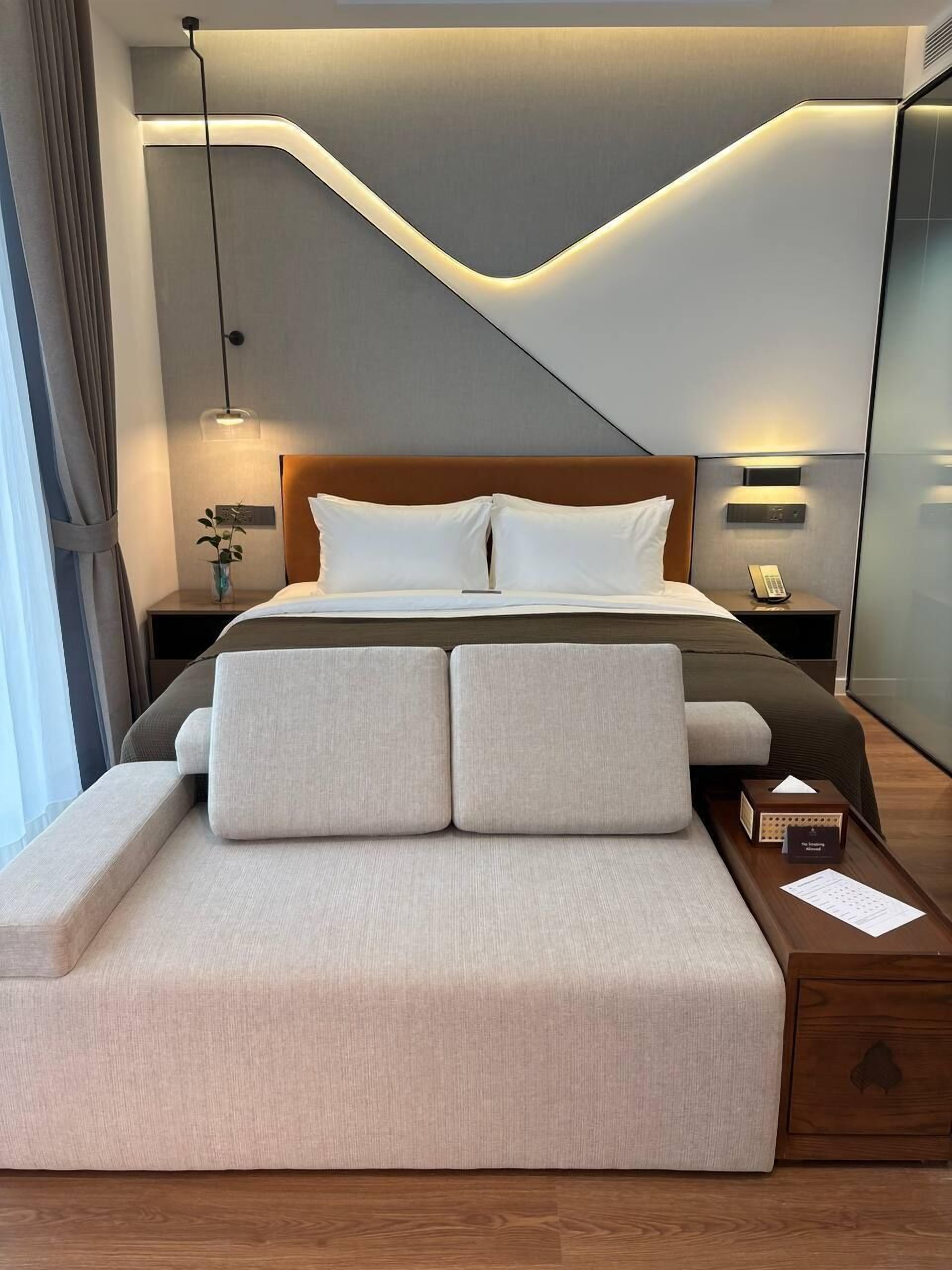 Une chambre moderne avec un plafond incurvé, une tête de lit en bois, une table de chevet avec une lampe et une petite table de nuit avec un téléphone.