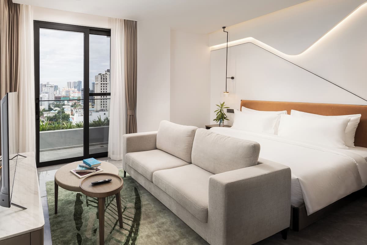 Quarto de hotel moderno com sofá, cama, uma mesinha com um controle remoto e vista da cidade através de uma grande janela.
