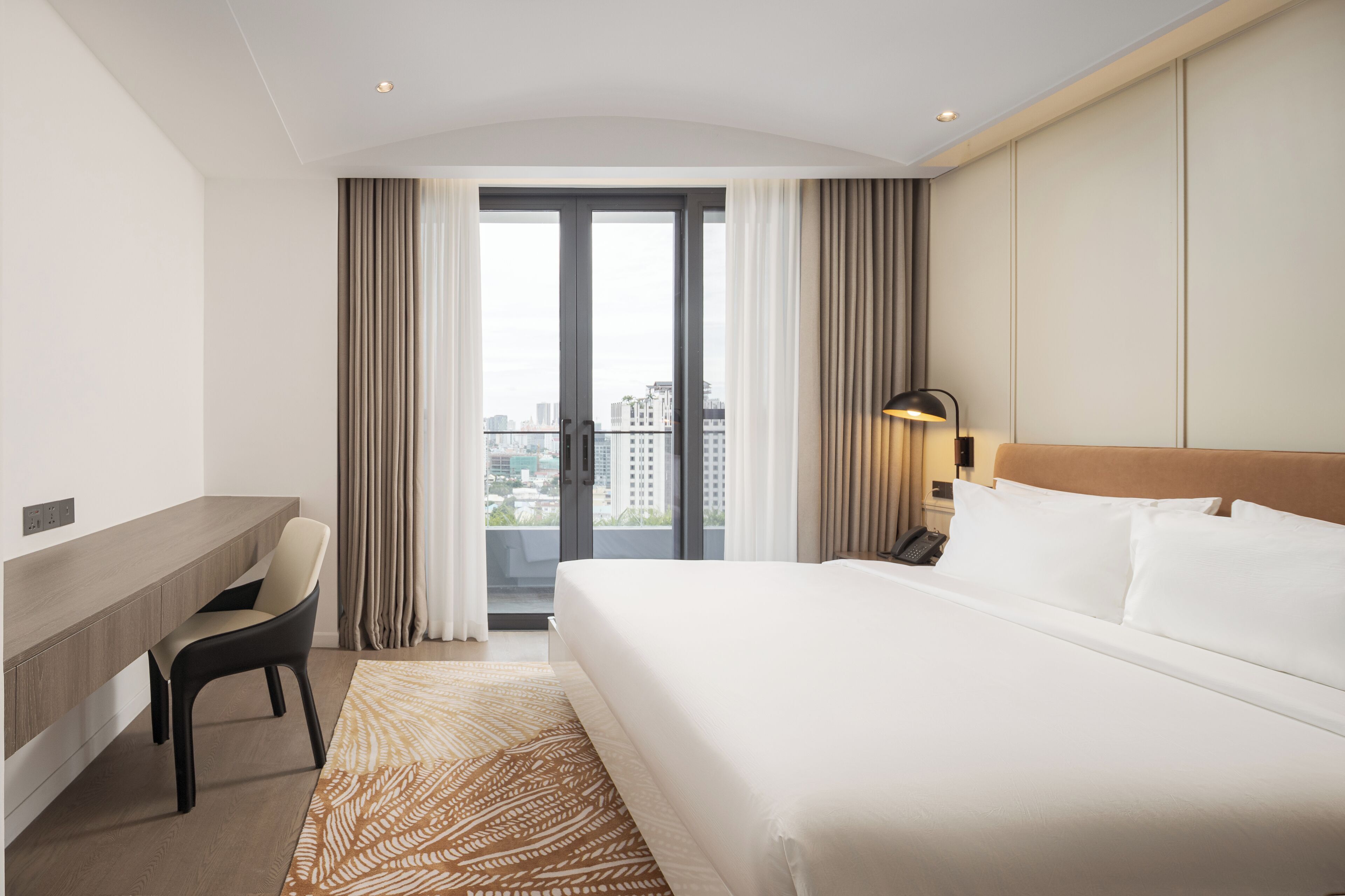 A hotel room with a bed, a desk, a chair, and a view of the city through the window.