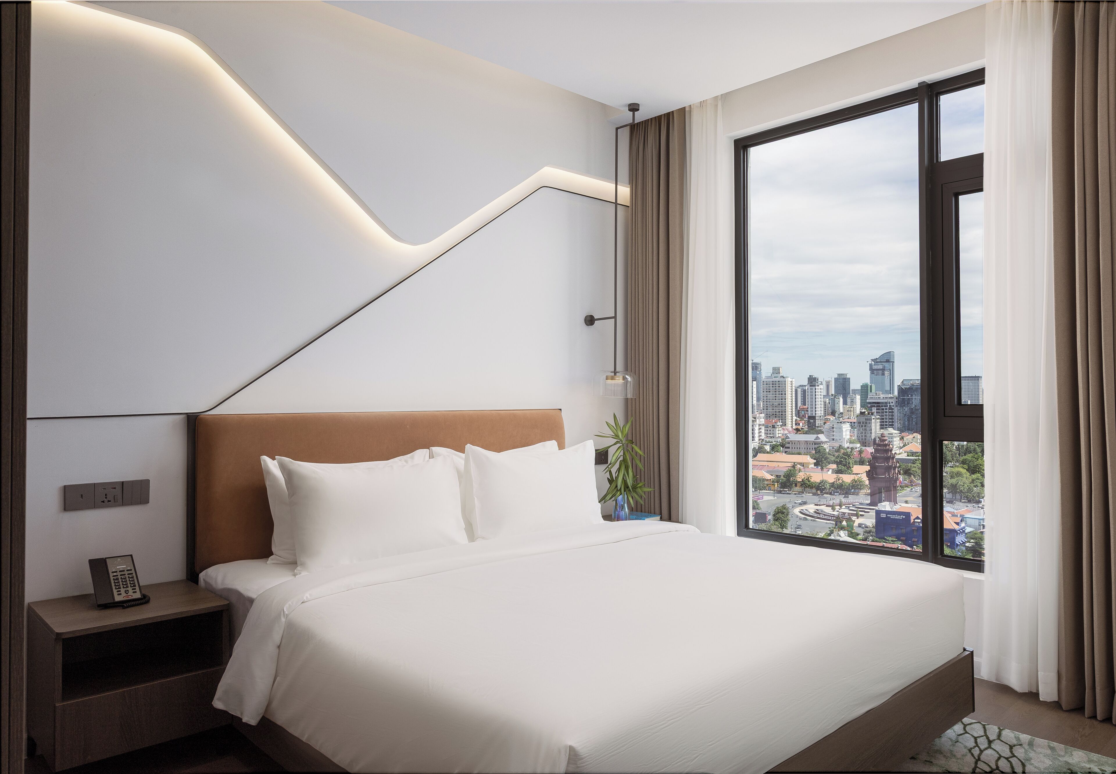 Ein modernes Hotelzimmer mit einem großen Bett, einem Nachttisch, einem Telefon und Blick auf die Stadt durch ein großes Fenster.