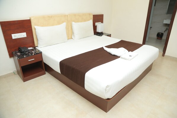 Cama bem arrumada com cabeceira marrom, almofadas brancas e lençol branco. Tem uma mesa de cabeceira com um telefone e uma luminária, e uma gavetinha.