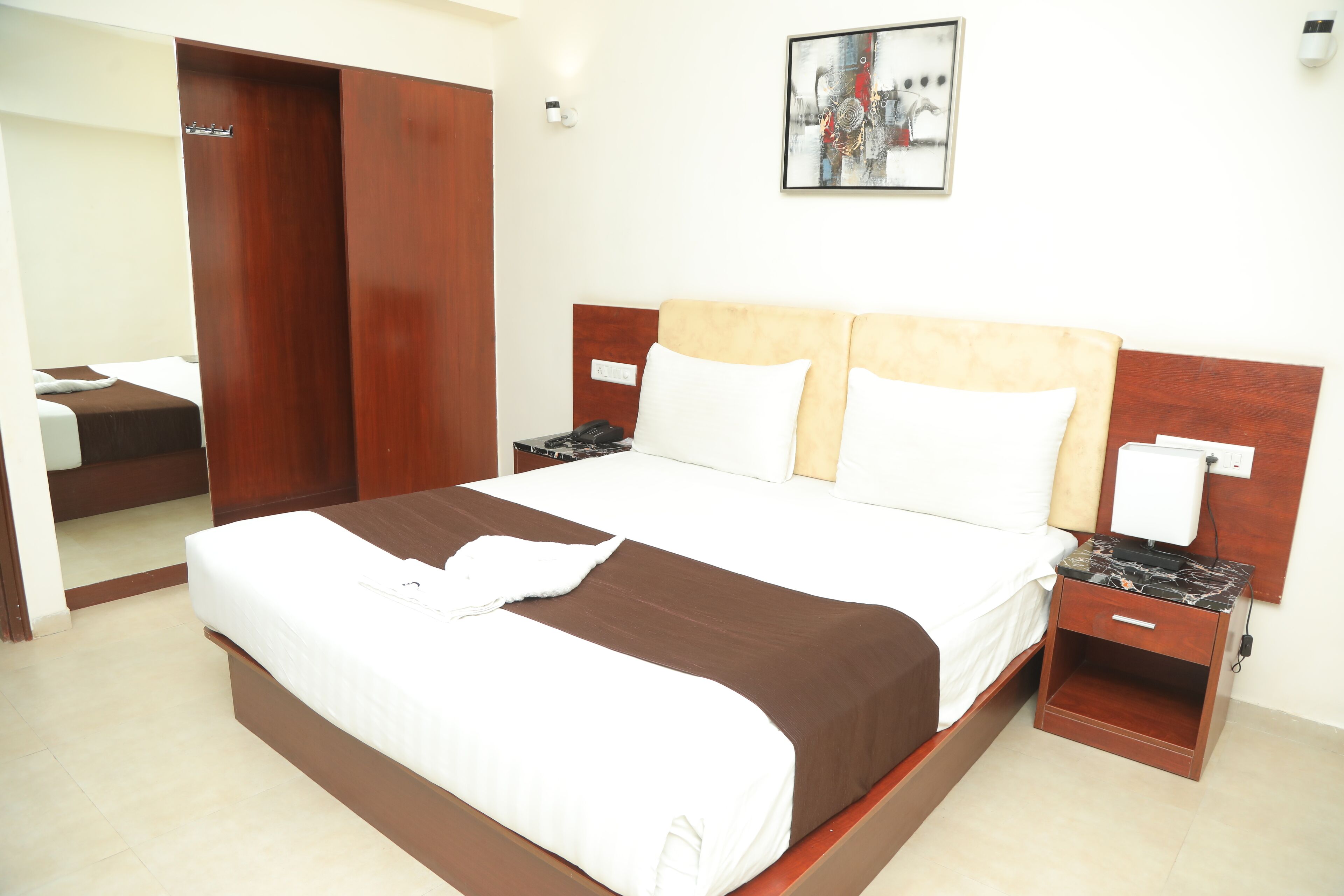 A hotel room with a large bed, wooden headboard, two bedside tables, a nightstand, a wardrobe, and a painting on the wall.