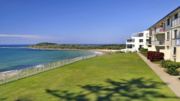 A coastal property with a grassy lawn, a white fence, and a building with balconies overlooking the sea.