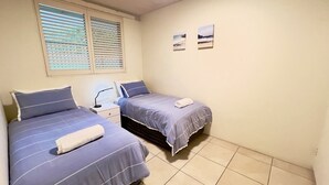 Ein Schlafzimmer mit zwei Betten, einem Fenster mit Jalousien, einem Nachttisch und zwei Wandbildern.