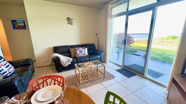 A living room with a black leather sofa, a glass door leading to a balcony, and a dining area with a table set for two.
