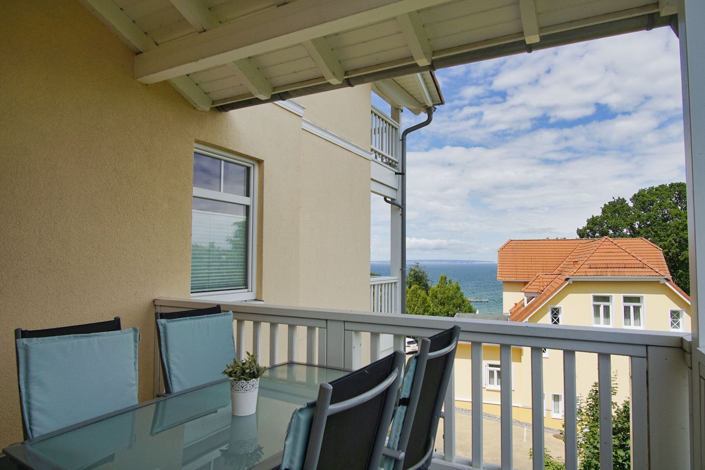 Uma varanda com mesa e cadeiras de vidro, um vaso de plantas e vista para o mar e edifícios.