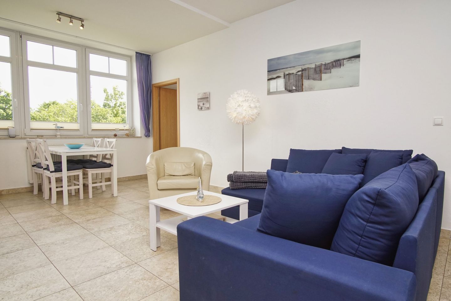 Una sala de estar con un sofá azul, un sillón color beige, una mesa de centro blanca y un comedor con mesa y sillas blancas.