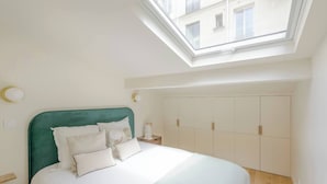A bedroom with a skylight, a bed with white bedding, a green headboard, and a wardrobe with multiple doors. - Apartment Como - Villa Alcôme (Paris)