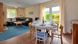 A dining area with a round table set for four, surrounded by wooden chairs. A sofa and a small coffee table are in the background. - Nant Y Mynydd (Amlwch)