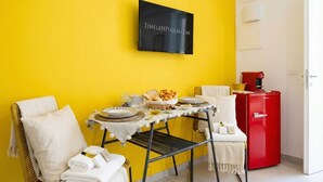 A bright yellow wall, a black sign with 'TIMELESSPUGLIA.COM', a table with plates and bread, a red fridge, and a white chair with a towel. - Dimora dei Colori - Yellow Light (Bari)