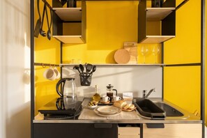 Et lille køkken med gul backsplash, en kaffemaskine, elkedel, et brød og et vinglas på bordpladen.