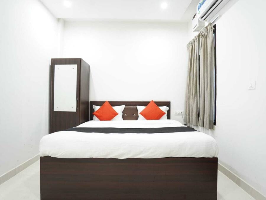 A modern hotel room with a large bed, dark wood headboard, white bedding, and two orange pillows.